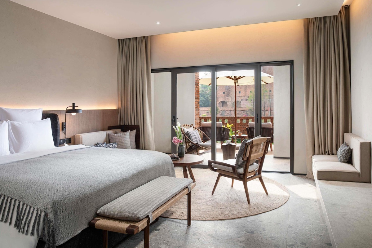 Una habitación de hotel moderna con una cama grande, un banco, una mesita pequeña y un balcón con sillas.
