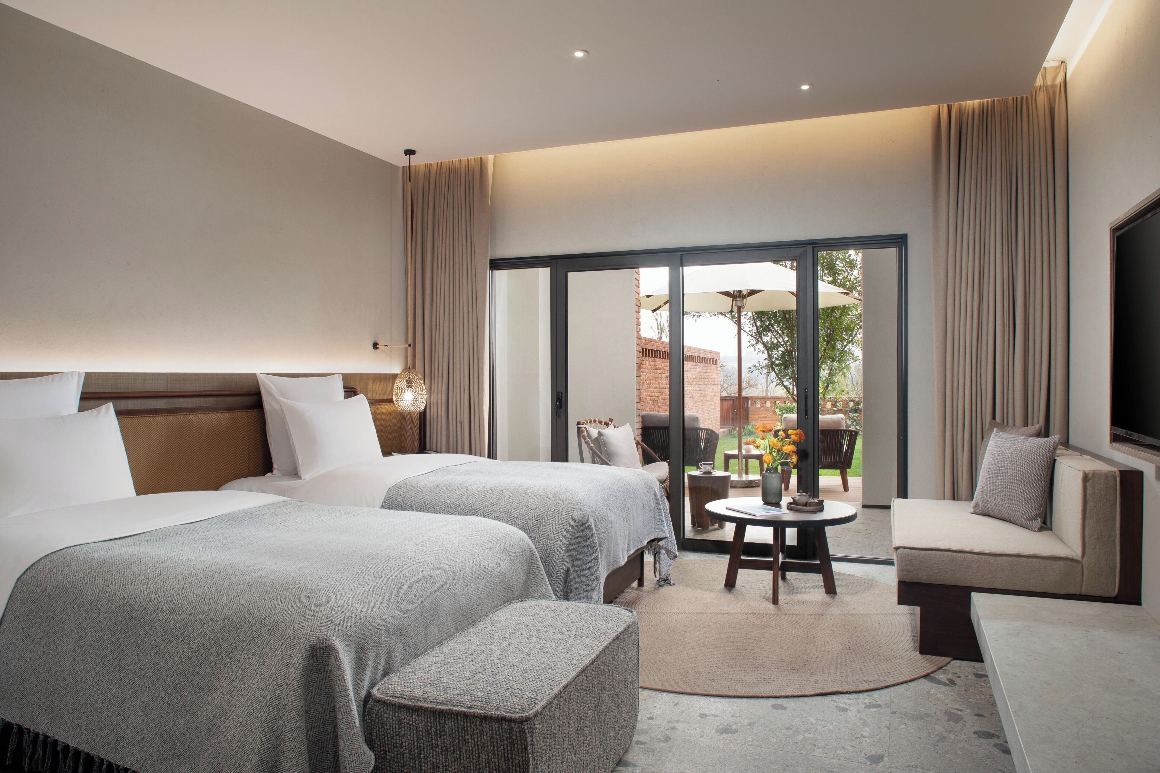 Habitación de hotel con dos camas, un sofá, un televisor y un ventanal corredero que da a un espacio exterior.