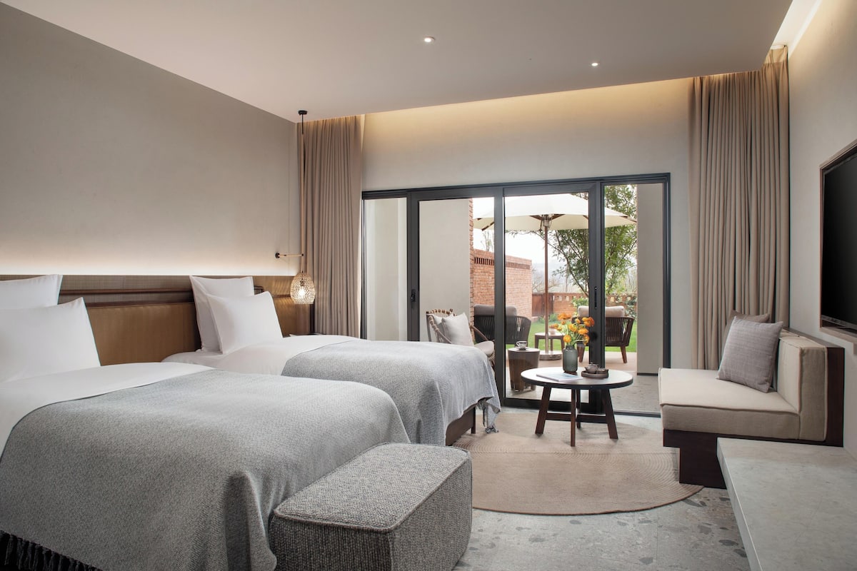 A hotel room with two beds, a sofa, a TV, and a sliding glass door leading to an outdoor area.