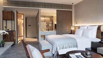 A modern hotel room with a large bed, a sitting area with a chair and table, a bathroom with a shower and mirror, and a wooden ceiling design.