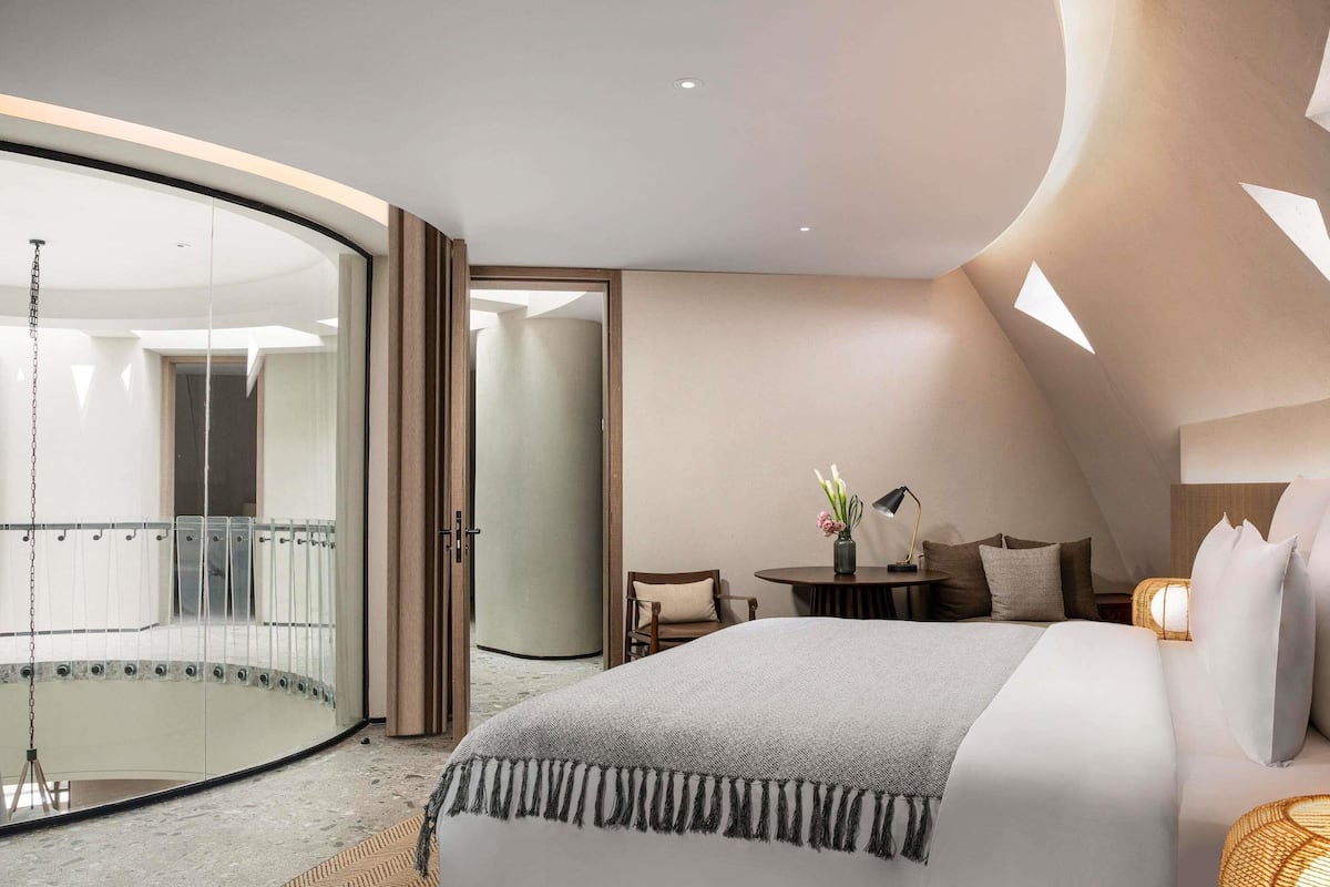 A modern bedroom with a curved ceiling, a large bed, a small table with a vase, and a balcony.