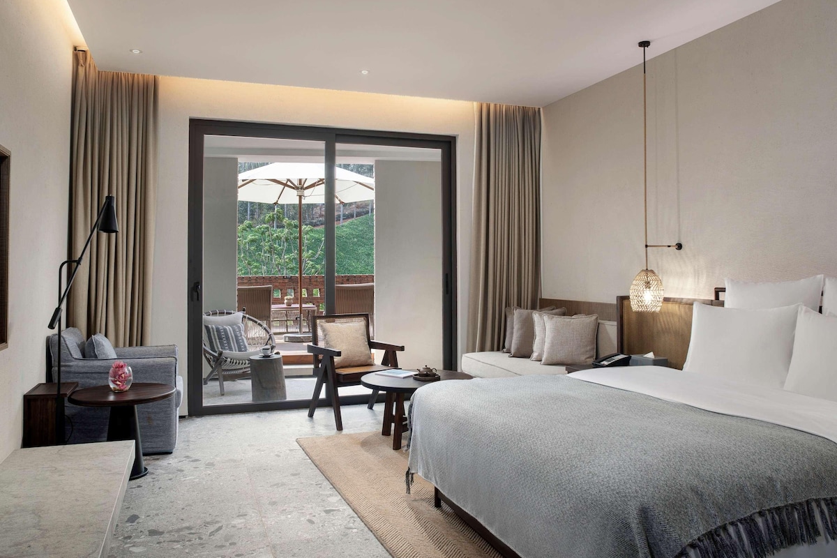 Habitación de hotel moderna con una cama grande, un sofá, una mesita y un balcón con vistas a la vegetación.