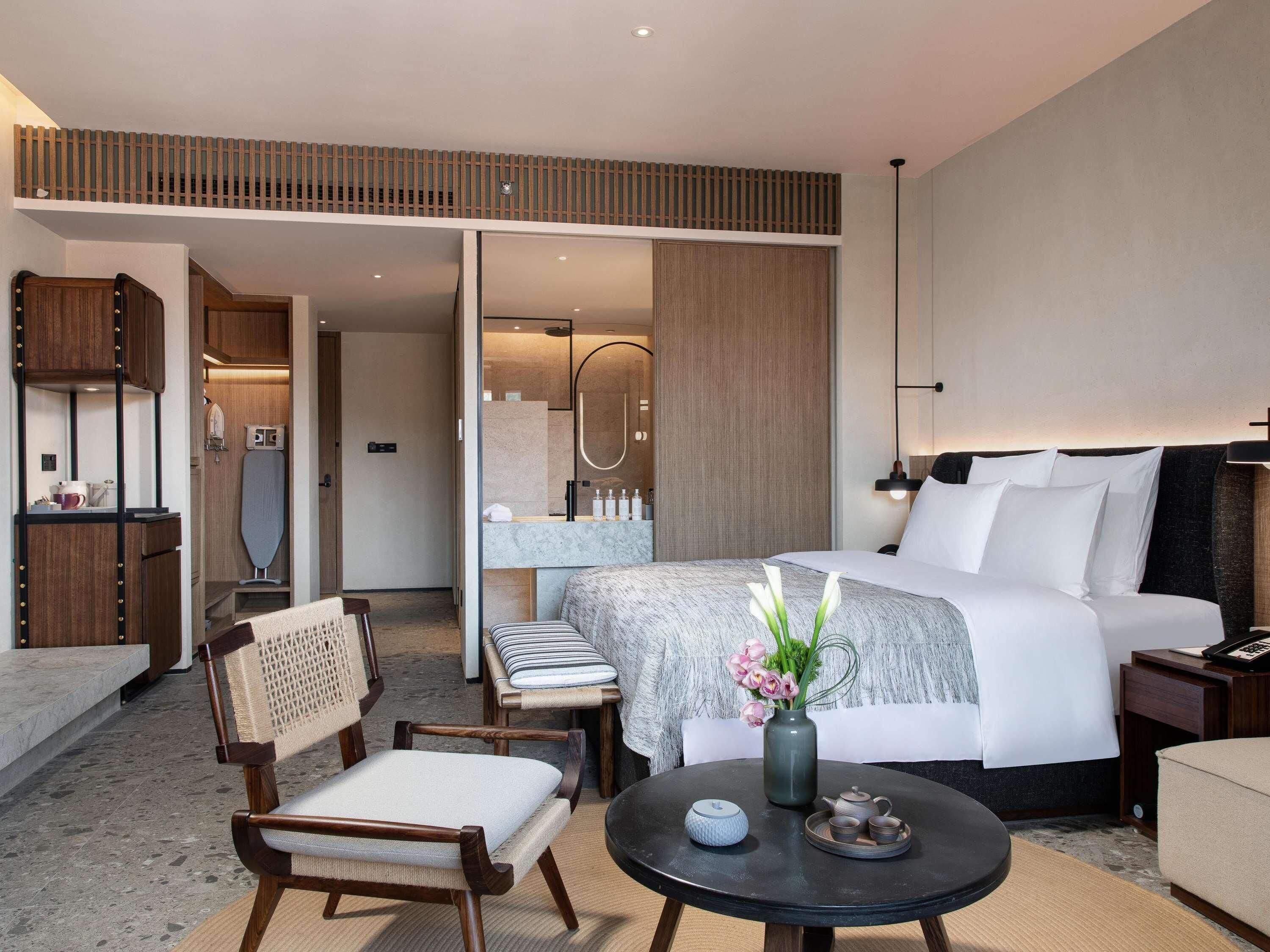 Een moderne hotelkamer met een bed, een bureau, een stoel, een ronde tafel met een vaas en theepot, en een badkamer zichtbaar door een geopende deur.