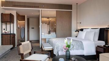 Een moderne hotelkamer met een bed, een bureau, een stoel, een ronde tafel met een vaas en theepot, en een badkamer zichtbaar door een geopende deur.
