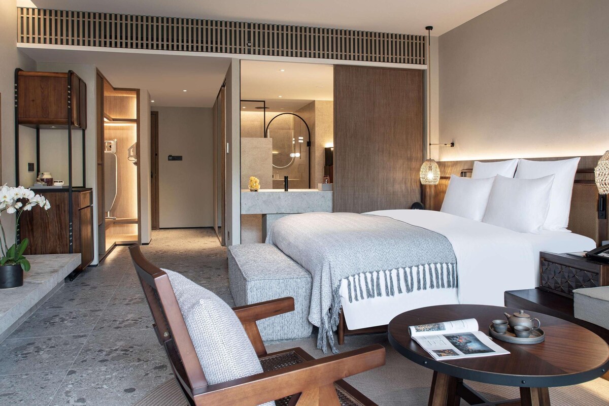 A modern hotel room with a large bed, a sitting area with a chair and table, a bathroom with a shower and mirror, and a wooden ceiling design.