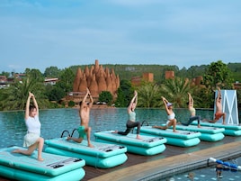 A group of people performing yoga on floating mats by a pool with a unique rock formation in the background.