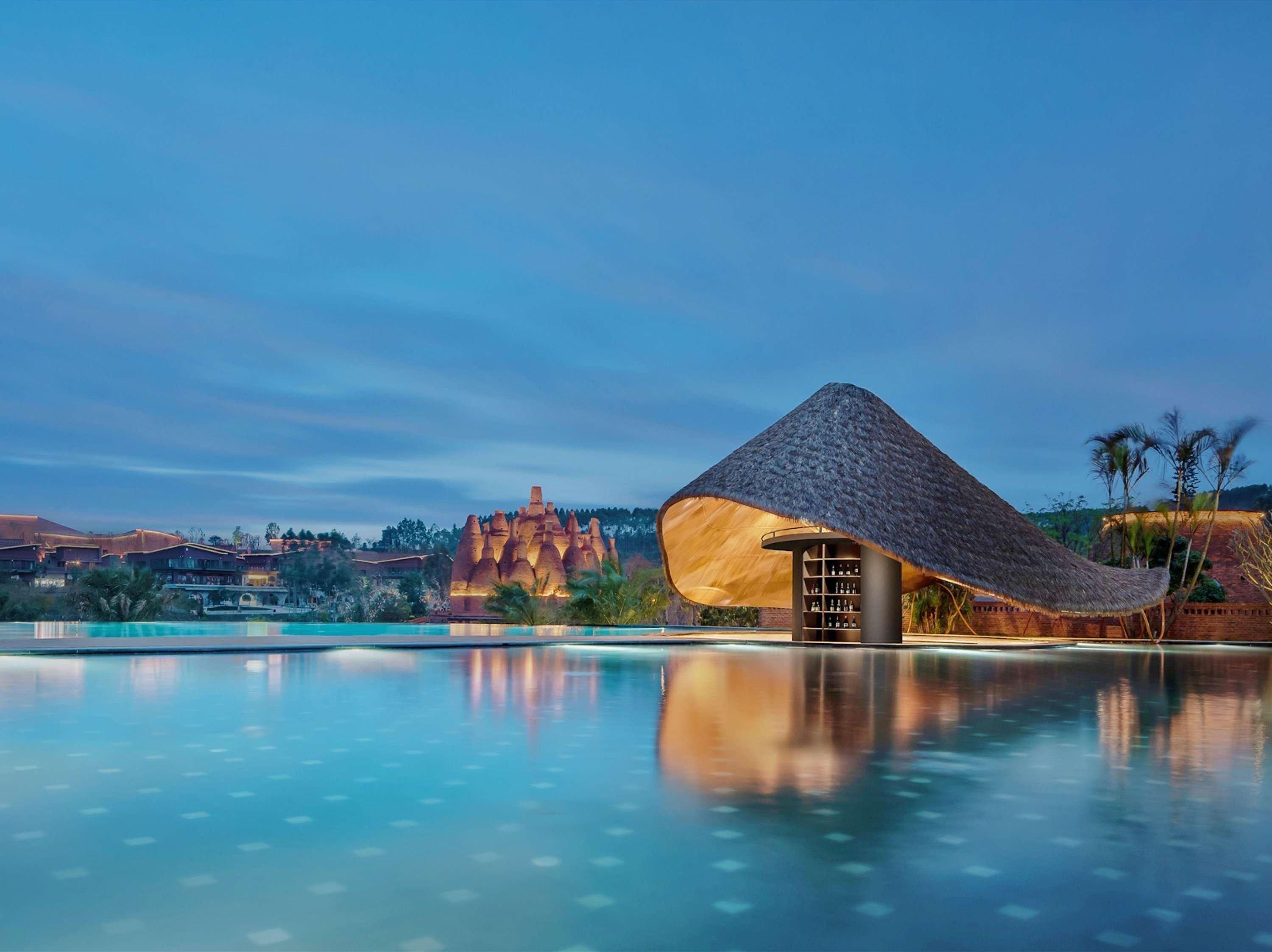 Un resort con piscina, una estructura de techo de paja y montañas al fondo.