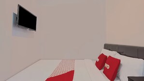 Una habitación de hotel moderna con un televisor de pantalla plana montado en la pared, una cama bien hecha con ropa de cama blanca y roja, y un cabecero gris.