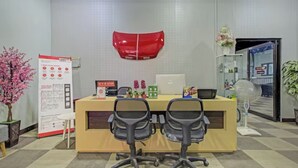 Un área de recepción de oficina con un escritorio, dos sillas, un adorno rojo en la pared, una pizarra y una planta en maceta.
