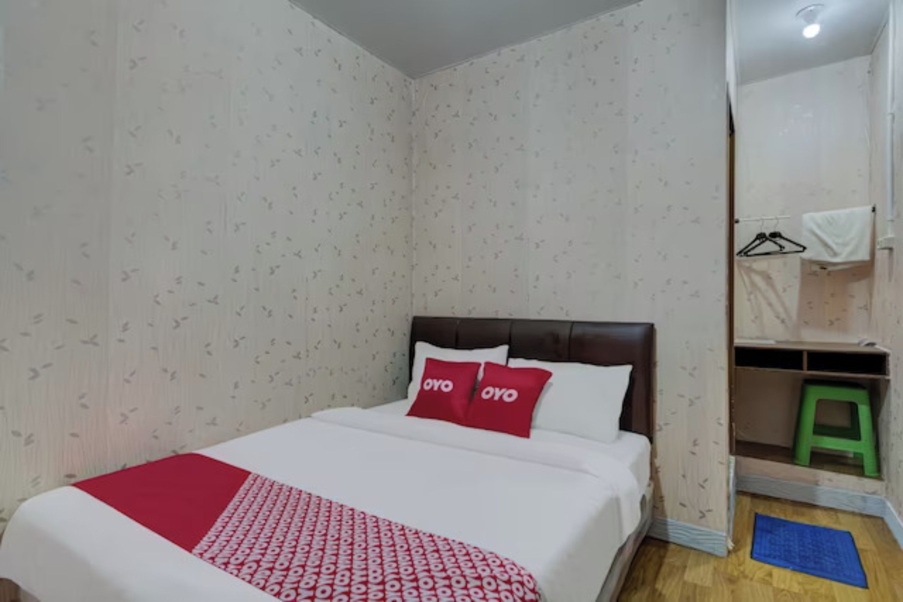 A bedroom with a bed, a red and white patterned bedspread, two red pillows, a wooden headboard, a green stool, and a blue mat on the floor.
