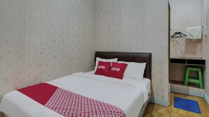 Un dormitorio con una cama, un edredón blanco y rojo estampado, dos almohadas rojas, un cabecero de madera, un taburete verde y un tapete azul en el suelo.