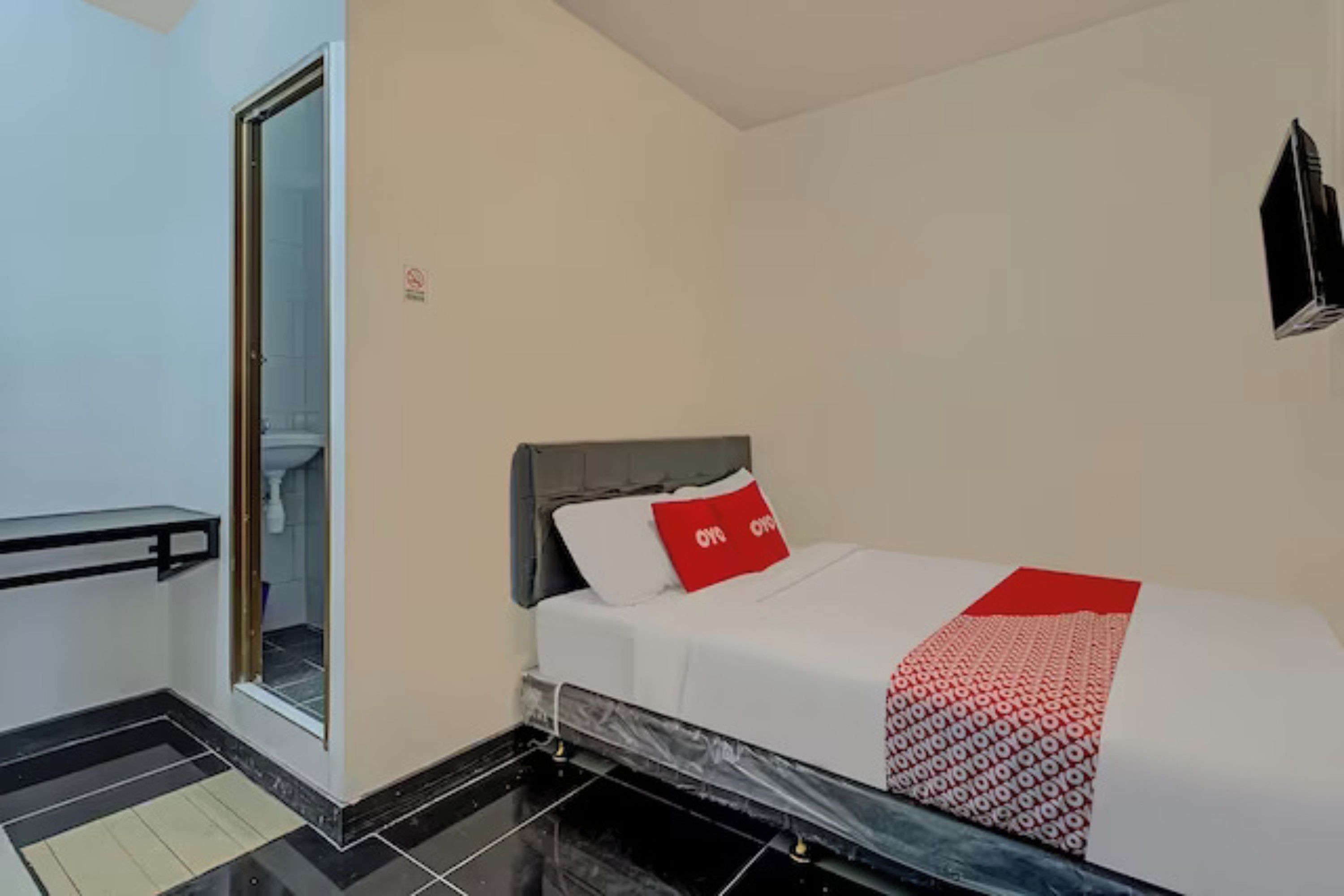 Un dormitorio con una cama, un almohadón rojo, una colcha blanca, una manta con estampado blanco y negro, un cabecero negro, una puerta de cristal que da a un baño y un televisor negro montado en la pared.