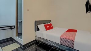 Un dormitorio con una cama, un almohadón rojo, una colcha blanca, una manta con estampado blanco y negro, un cabecero negro, una puerta de cristal que da a un baño y un televisor negro montado en la pared.