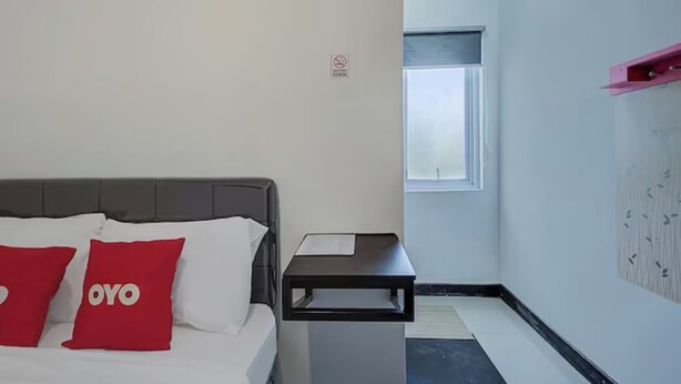 Habitación de hotel con cama, dos almohadas rojas, una mesita de noche, una ventana y un estante en la pared.