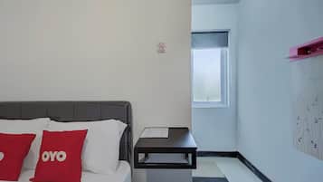 A hotel room with a bed, two red pillows, a small bedside table, a window, and a wall-mounted shelf.