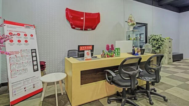 Una recepción con un escritorio, una computadora, un teléfono y una pequeña planta. Hay un cartel en la pared y una silla frente al mostrador.