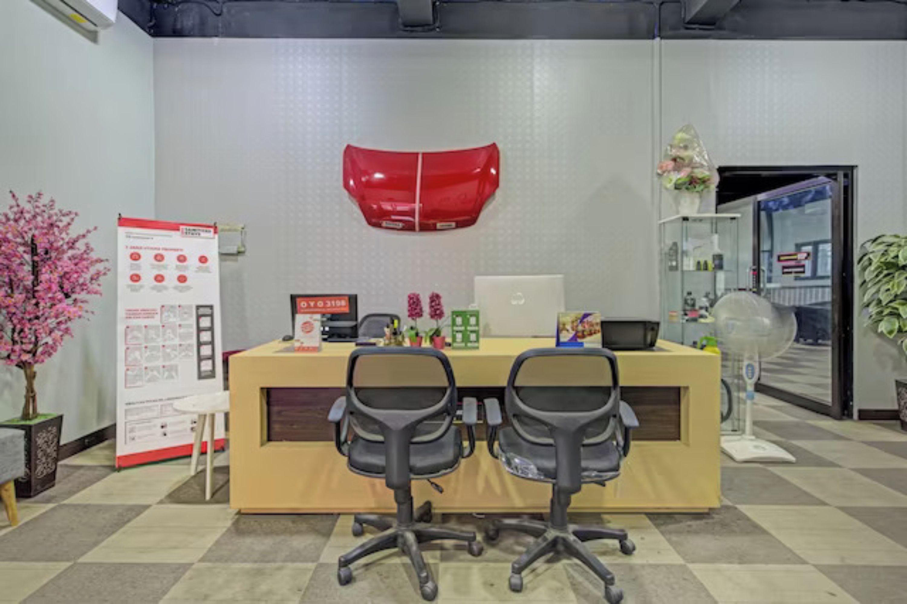 Un área de recepción de oficina con un escritorio, dos sillas, un adorno rojo en la pared, una pizarra y una planta en maceta.