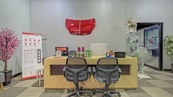 Un área de recepción de oficina con un escritorio, dos sillas, un adorno rojo en la pared, una pizarra y una planta en maceta.