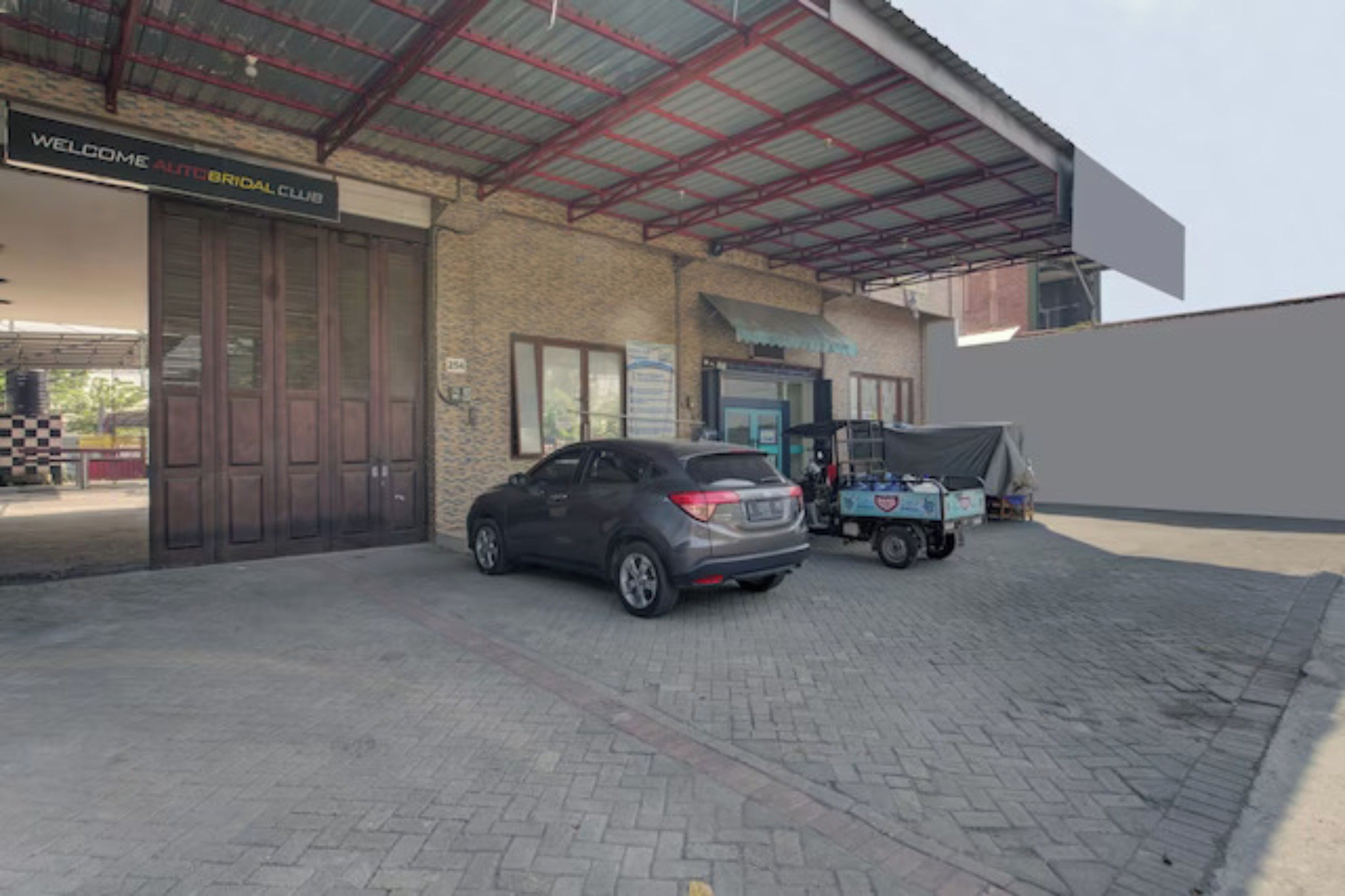 A building with a sign reading 'Welcome Auto Bridal Club,' a parked car, and a covered area with a blue vehicle.
