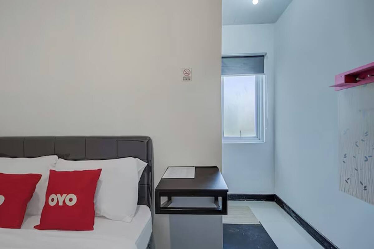 Quarto de hotel com uma cama, duas almofadas vermelhas, uma mesa de cabeceira pequena, uma janela e uma prateleira fixada na parede.