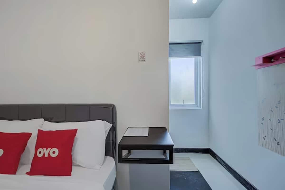 Habitación de hotel con cama, dos almohadas rojas, una mesita de noche, una ventana y un estante en la pared.