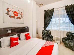 A hotel room with a bed, two chairs, a window with curtains, and a wall with a framed picture.