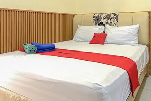 Un letto rifatto con biancheria bianca e una coperta rossa, un cuscino con il logo rosso, e un asciugamano blu.