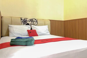 Una camera d'albergo con un letto, cuscini, una coperta rossa e una borsa verde.