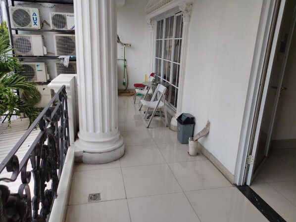 Un balcon propre et bien entretenu, avec des colonnes blanches, un sol carrelé et une vue sur l’extérieur. On y trouve, à gauche, des unités de climatisation, une échelle et, à droite, une poubelle.