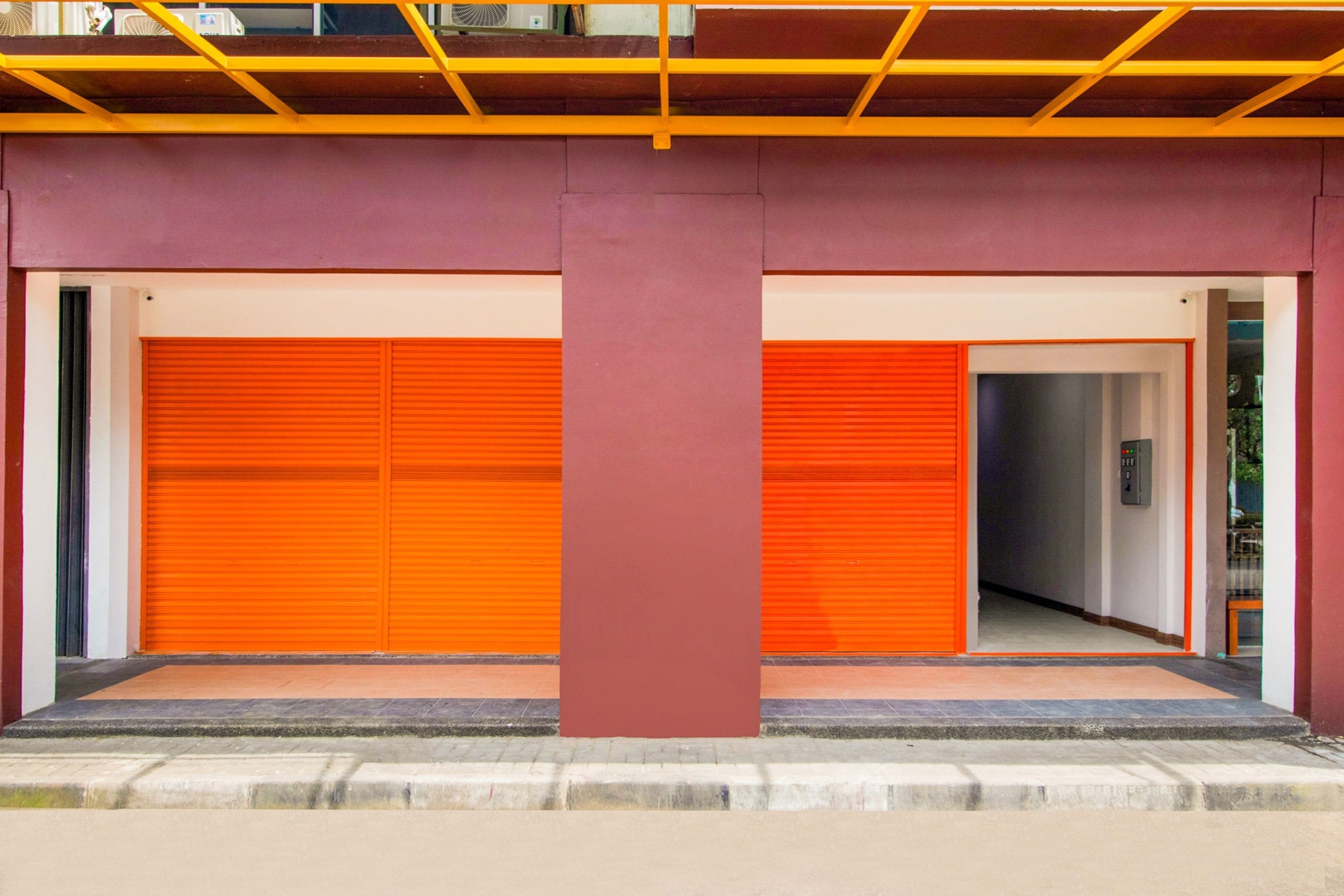 A building with two closed orange garage doors, a central red pillar, and a concrete ground level.
