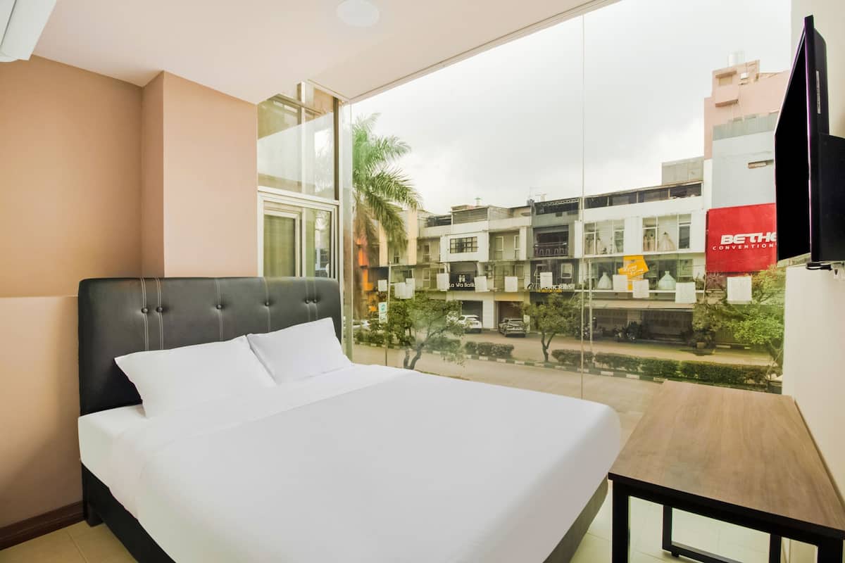 A hotel room with a large bed, a wooden nightstand, and a view of the city through a large window.