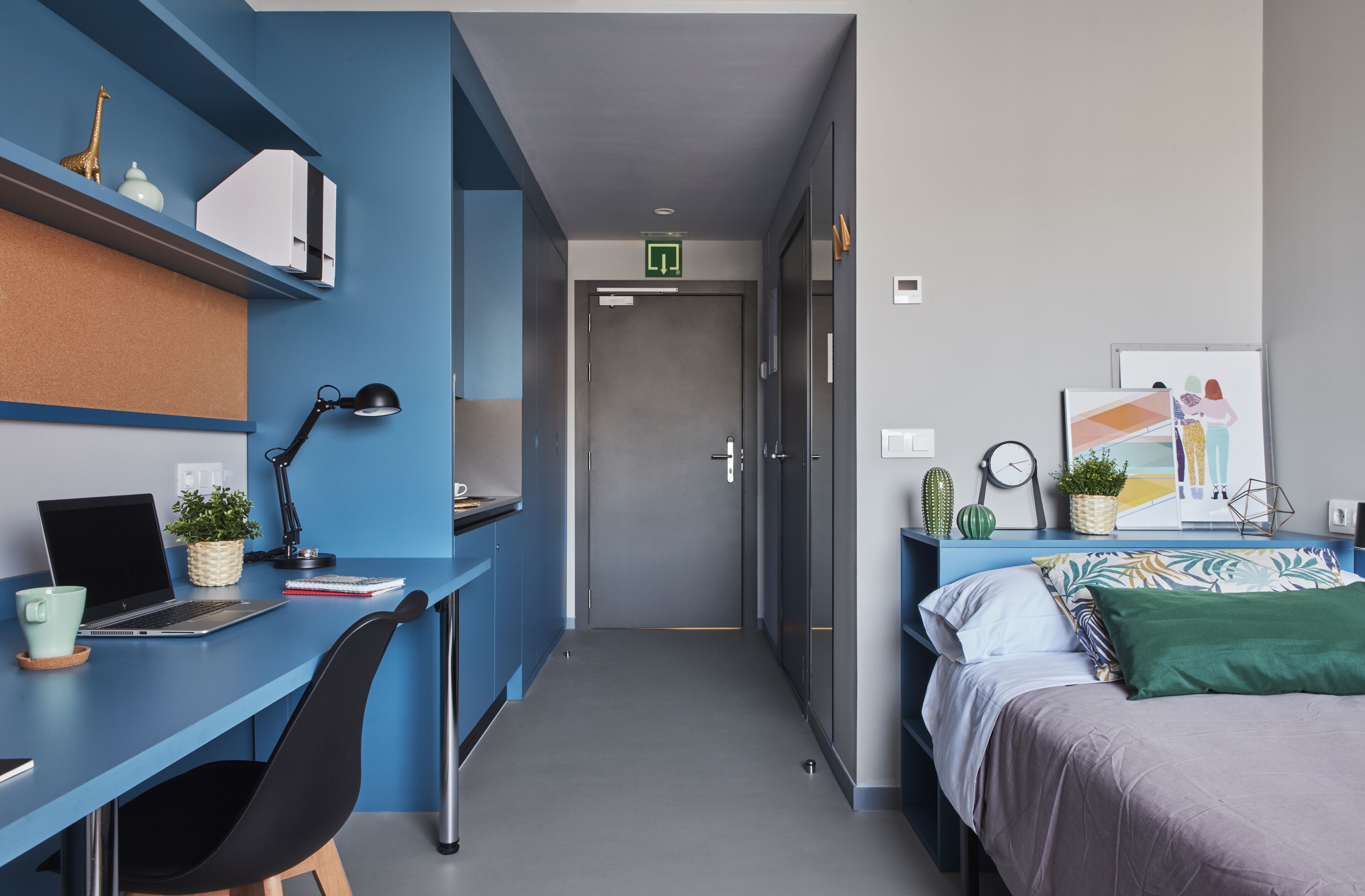 A modern hotel room with a blue desk, a laptop, a chair, a bed with a green pillow, and a small plant.