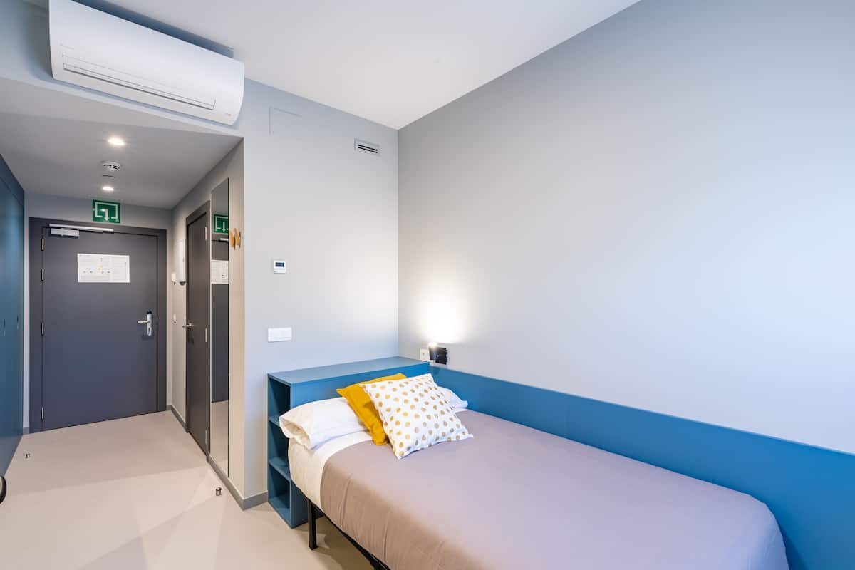 Een slaapkamer met een bed, een nachtkastje, een wandlamp en een deur met een nooduitgangsbord.
