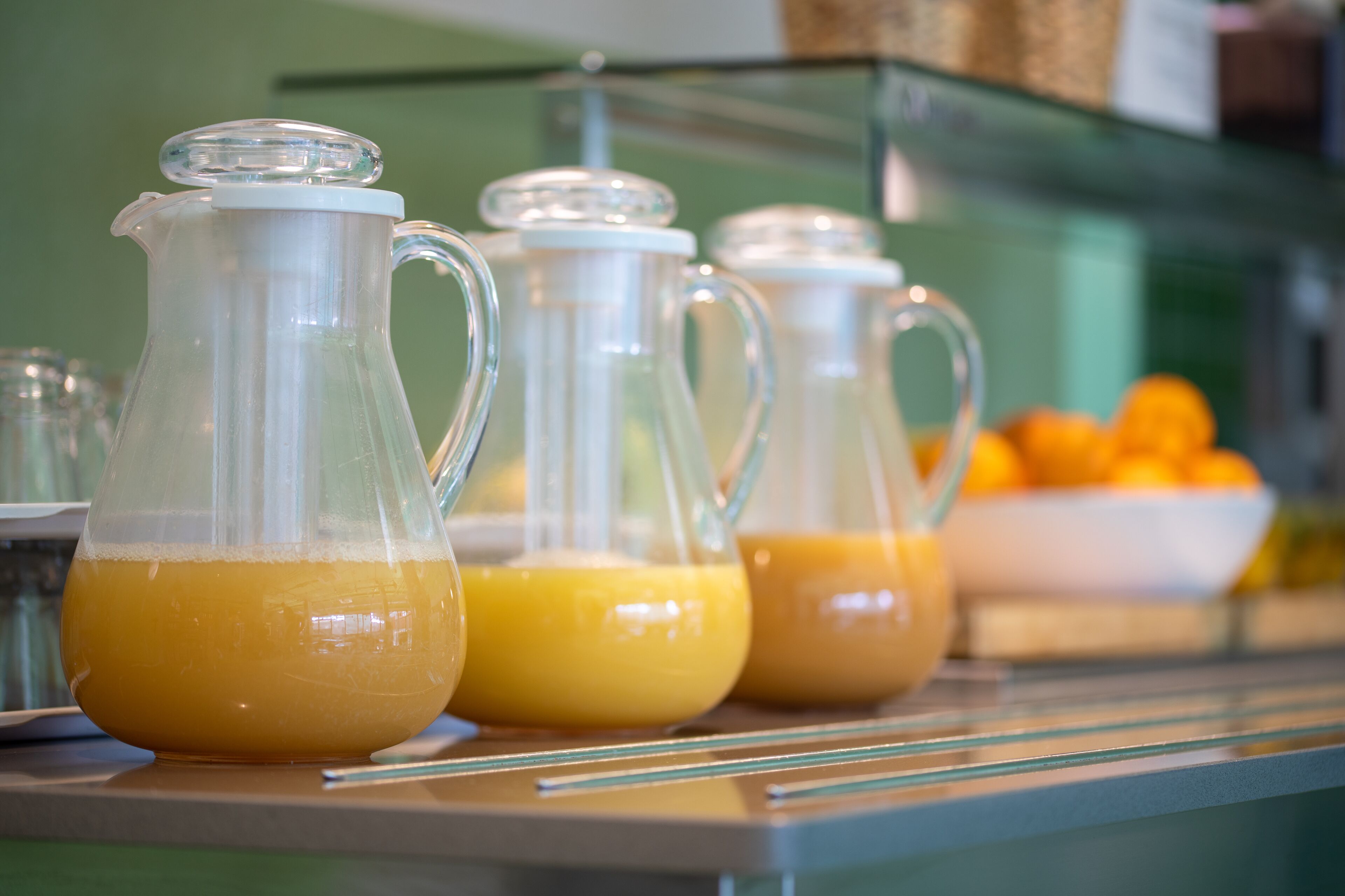 Three glass pitchers of orange juice on a counter with a bowl of oranges in the background.