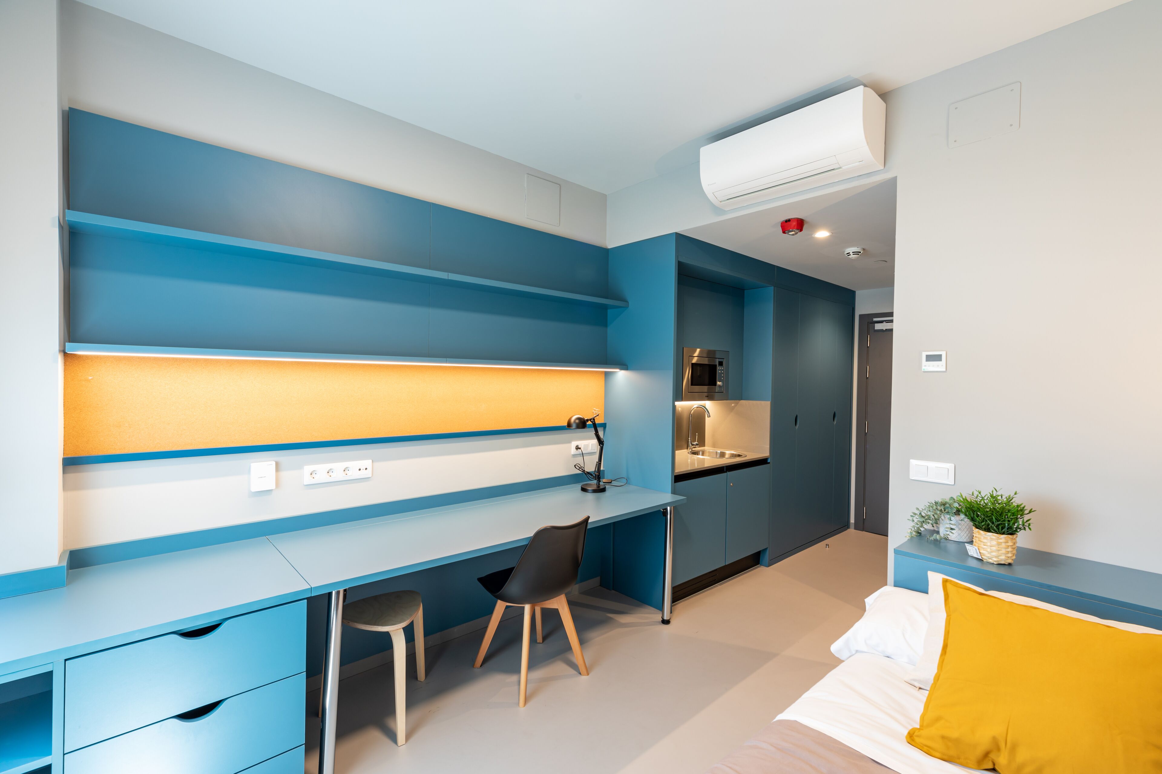 A modern room with a blue and white color scheme, featuring a desk, chair, bed, and a small kitchenette.