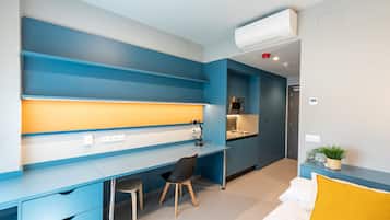 A modern room with a blue and white color scheme, featuring a desk, chair, bed, and a small kitchenette.