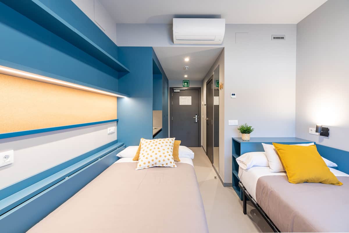 Een hotelkamer met twee bedden, een blauwe accentmuur, een kleine plant en een luchtverfrisser aan de muur.