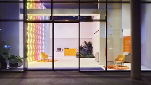 Un edificio moderno con amplios ventanales, una recepción con un mostrador amarillo y dos sillas amarillas en el exterior.