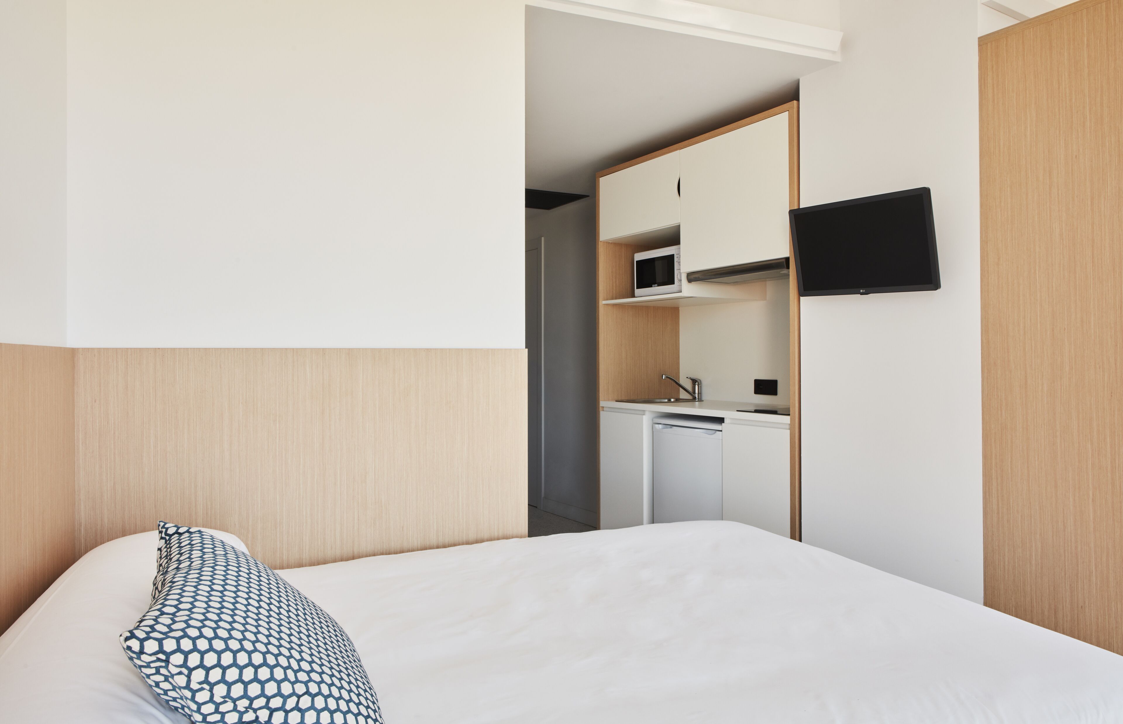 A hotel room with a bed, a patterned pillow, a television mounted on the wall, and a kitchenette area with a microwave and sink.