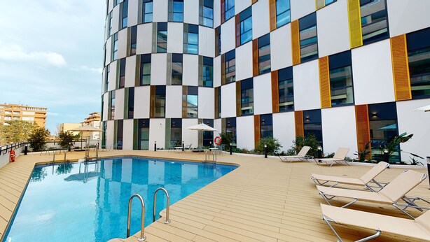 Un edificio moderno con un área de piscina que incluye sillas de descanso y una piscina de color azul claro.
