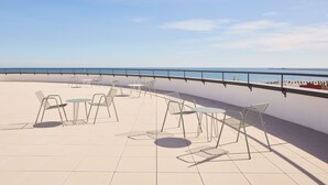 Terraza exterior con sillas y mesas de metal, con vistas a un cuerpo de agua y un barco a lo lejos.