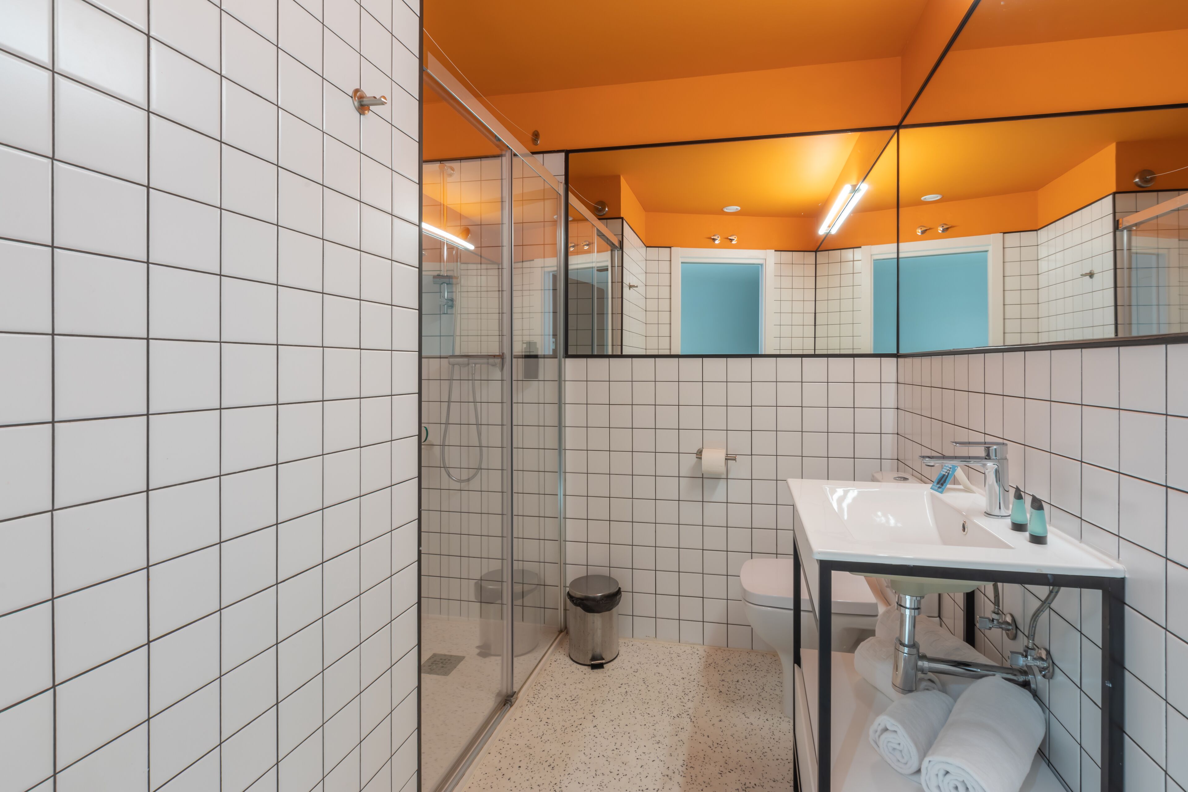 A modern bathroom with white tiled walls, a glass shower enclosure, a white sink with a mirror, and a blue toilet.