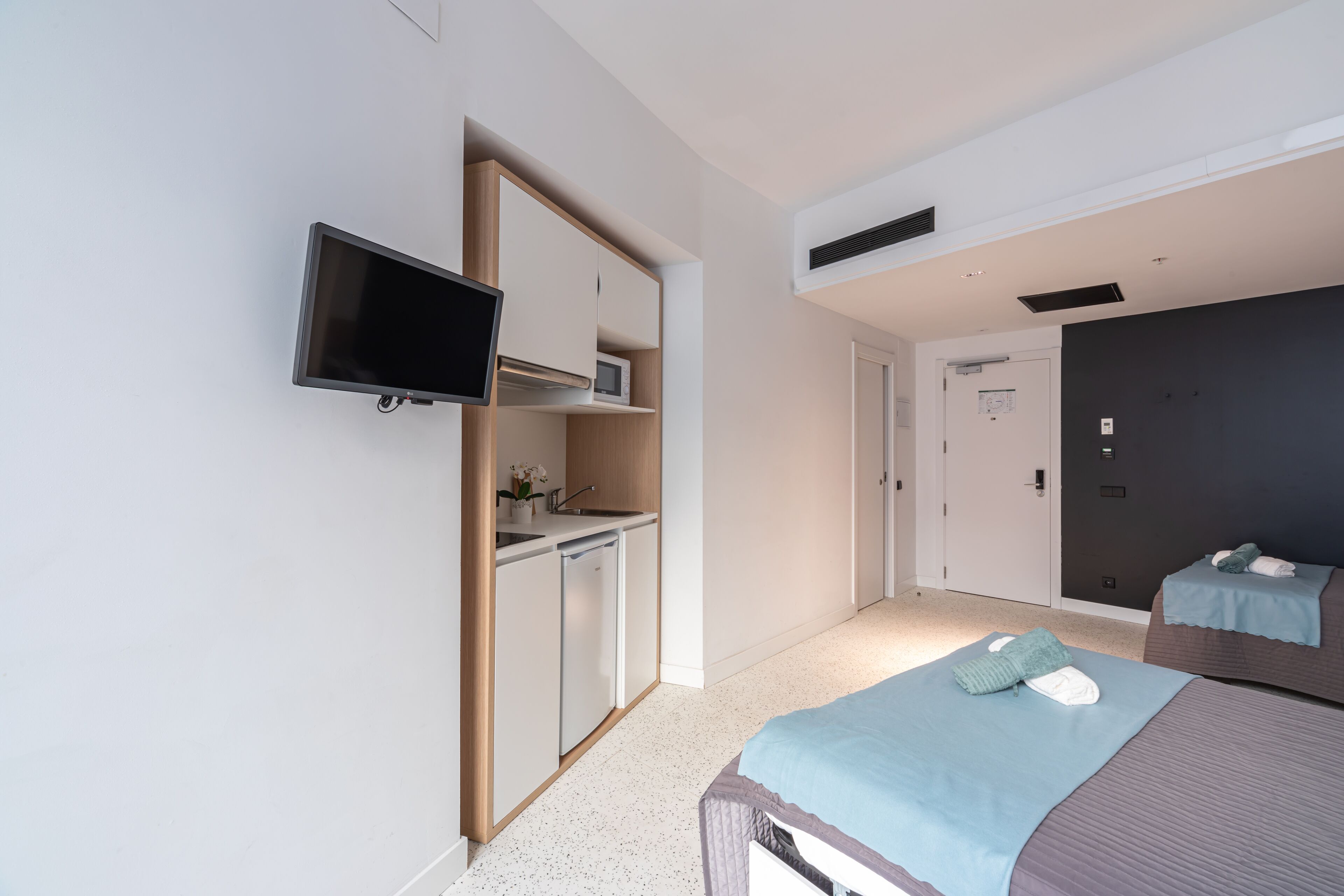 A modern hotel room with a kitchenette, a flat-screen TV, a bed with a blue and brown bedspread, and a dark-colored wall.