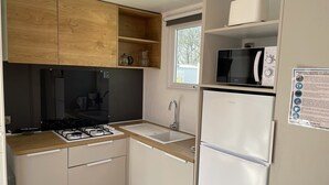 A compact kitchen with a gas stove, sink, and white refrigerator. There is a window above the sink, and a small shelf with kitchen items. - MOBILE HOME R10 ALL COMFORT - 5* CAMPSITE (Quimper)