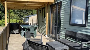 A covered outdoor patio with a wooden deck, a table with chairs, and a sliding glass door leading to a house. - MOBILE HOME R10 ALL COMFORT - 5* CAMPSITE (Quimper)