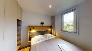 A modern bedroom with a wooden headboard, a large window, and built-in shelving. - MOBILE HOME R10 ALL COMFORT - 5* CAMPSITE (Quimper)