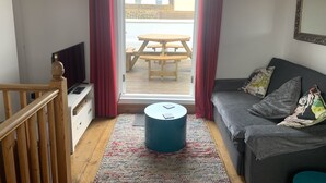 A room with a wooden floor, a grey sofa with colorful cushions, a small blue ottoman, a wooden table and chairs outside a sliding glass door, and a TV on a wooden stand. - Laines Loft - cosy flat in the centre of The Laines, with private sun terrace (Brighton)