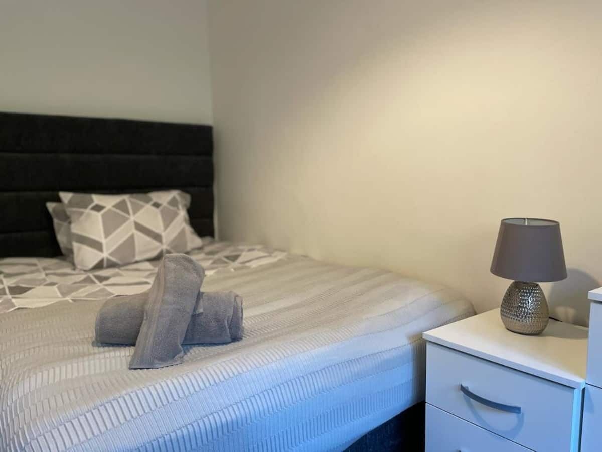 Ein ordentlich bezogenes Bett mit gestreifter Tagesdecke und zwei Kissen, eine Nachttischlampe auf einem Nachttisch und eine weiße Kommode.