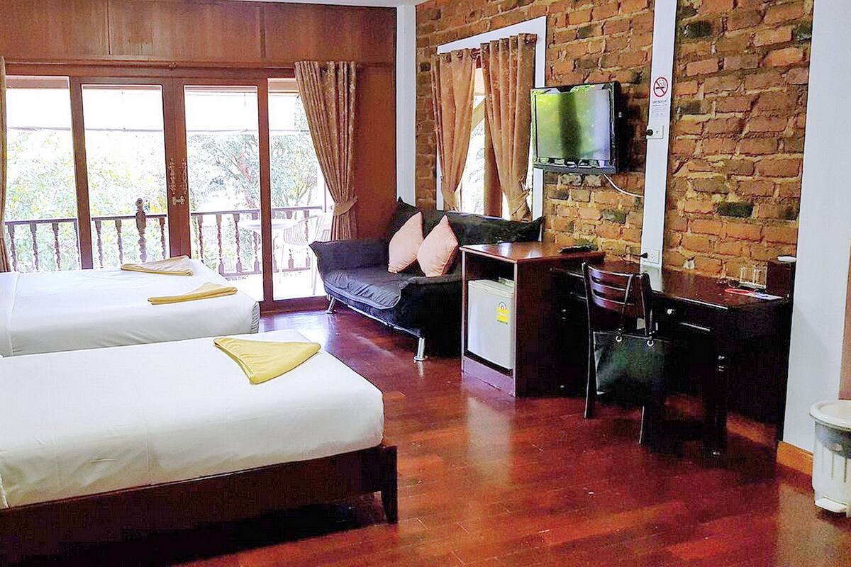 A hotel room with two beds, a sofa, a desk, and a TV mounted on a brick wall.