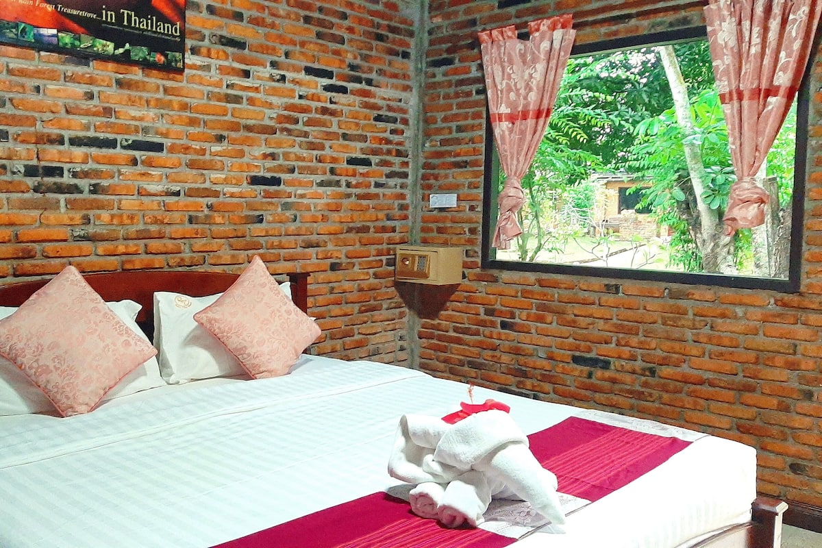 A room with a brick wall, a bed with white bedding and pink pillows, a window with a view of greenery, and a sign on the wall.